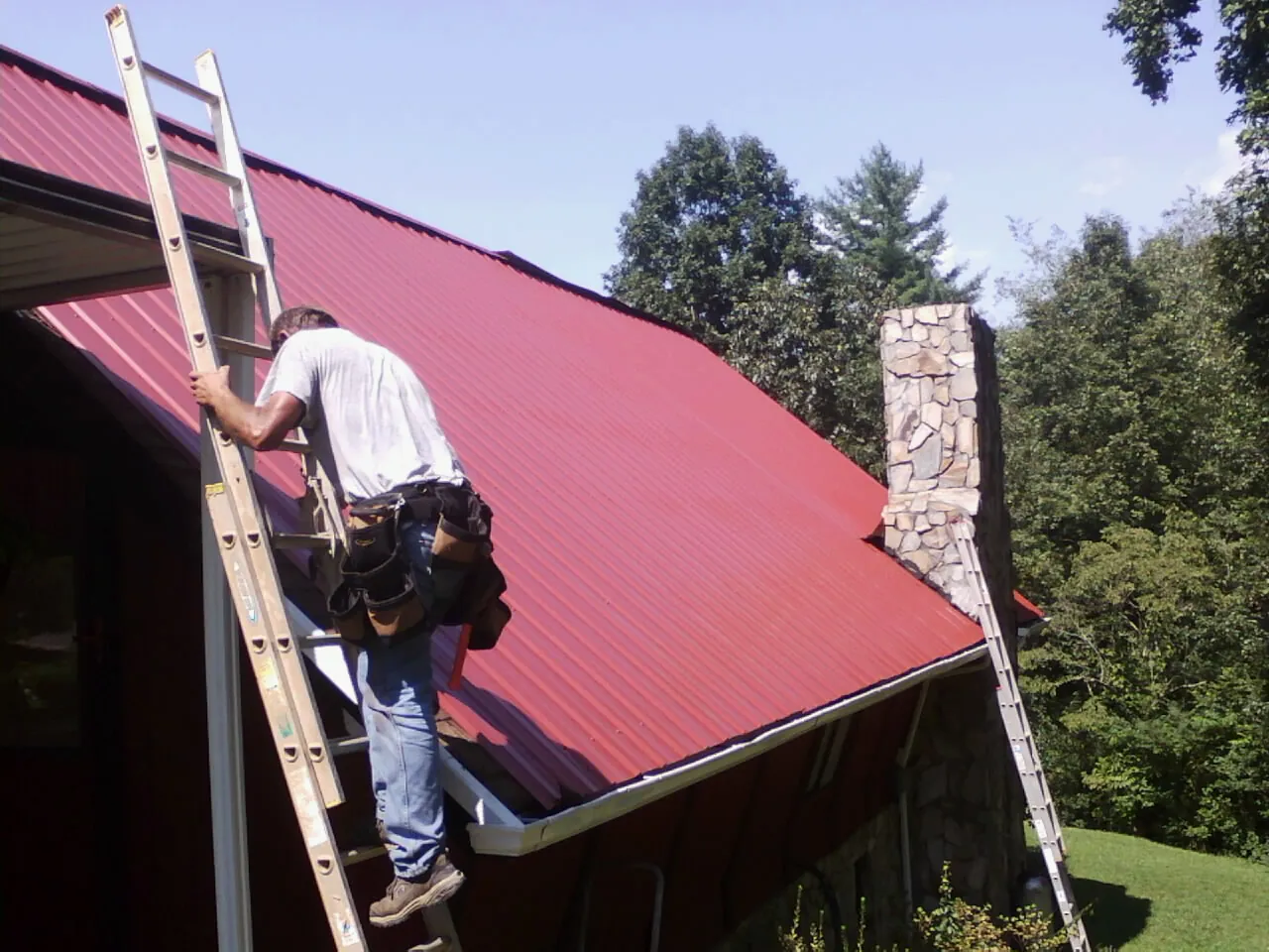 Roofer on a ladder beside a red metal roof and stone chimney — Soffit & Fascia Repair by Roofing Brothers in Shelton
